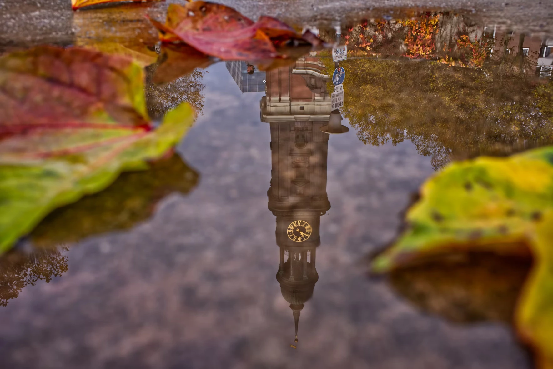 A puddle on a footpath with the reflection of a clock tower in it.